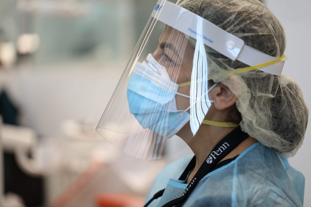 A dentist wears PPE with her hair in a net & waits for her next patient at a Penn Dental Medicine student dental care clinic.
