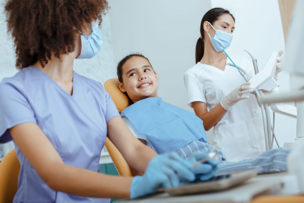  Masked student dentists at dental school that takes patients prepare to examine and clean teeth of girl in dental chair. 