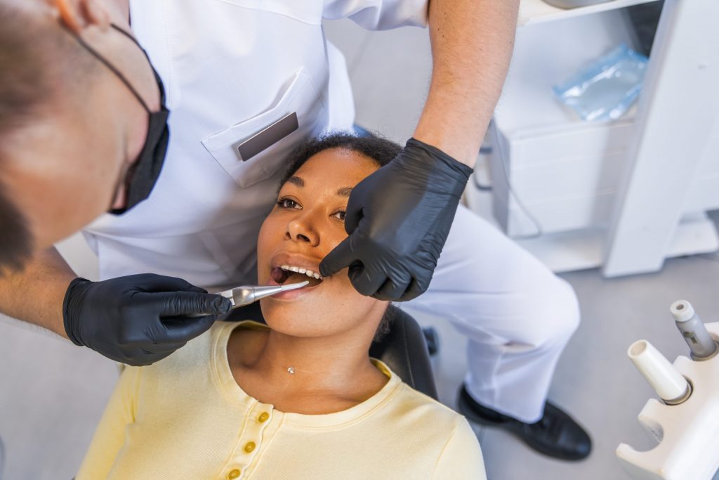 Masked and gloved dentist holds dental curing light in mouth of a woman reclined in dental clinic chair to harden a filling.