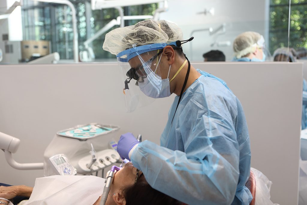  Dentist wearing mask, face shield, gloves, and scrubs examines teeth of man reclining in chair for dental treatment. 