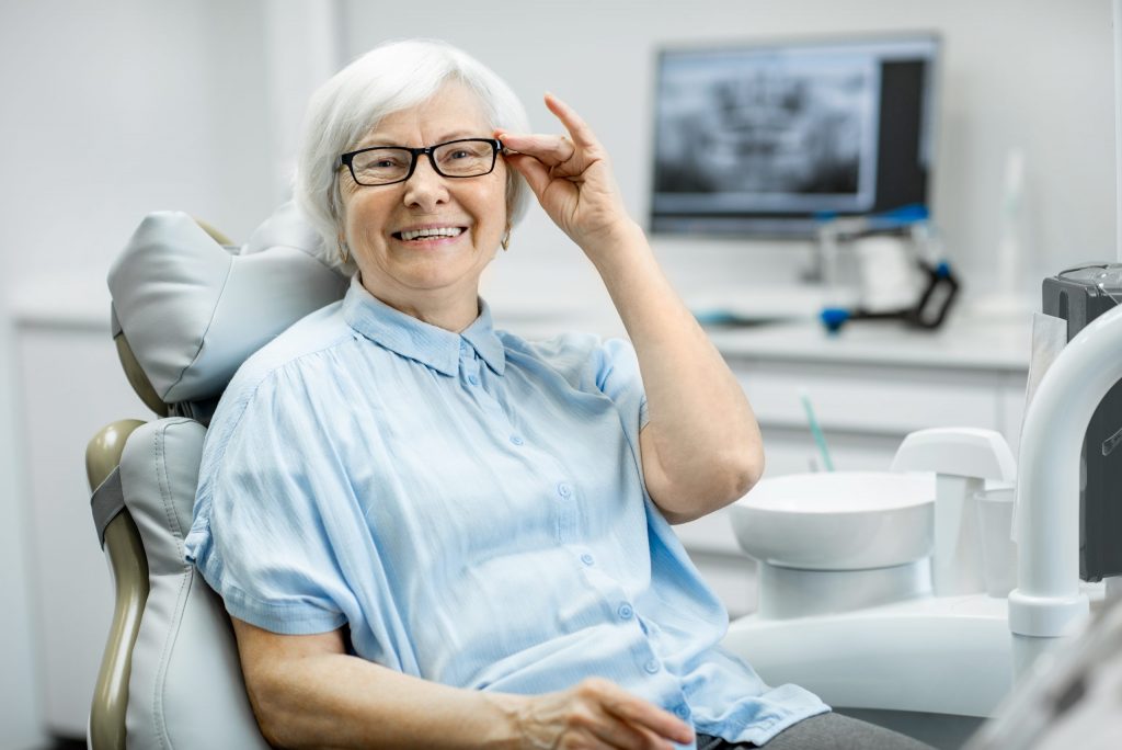  Woman sits in dental chair, adjusting her glasses and smiling after dental treatment at low-income dental clinic. 
