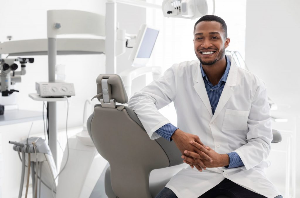  Student dentist who provides affordable dental care, wears a medical coat, and smiles as he poses in his office’s dental chair. 