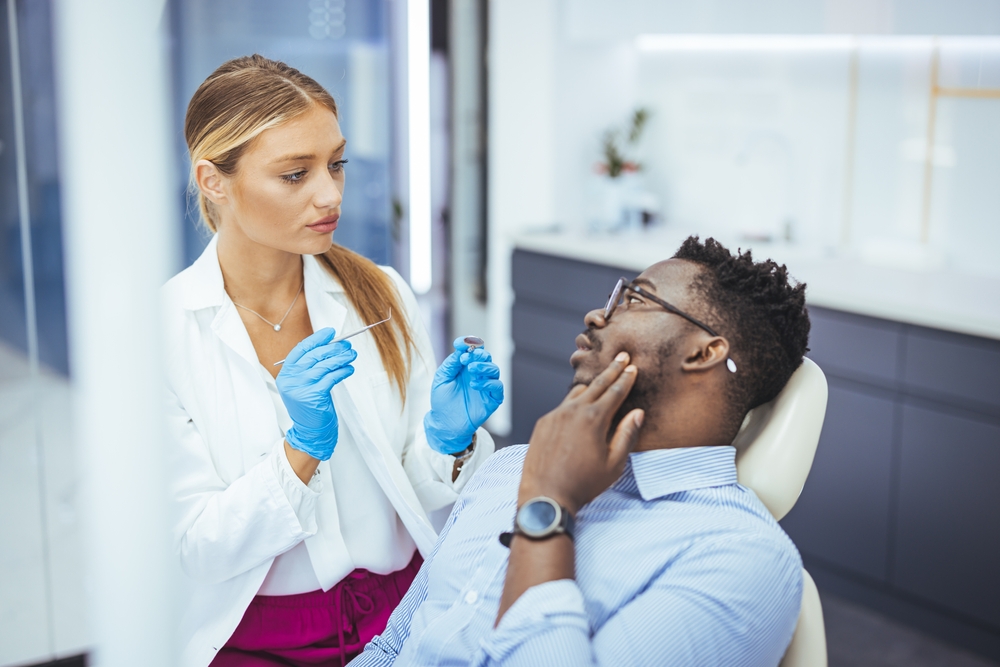 Philadelphia endodontist listens to her patient in dental chair, who holds his left hand to cheek to indicate dental pain.