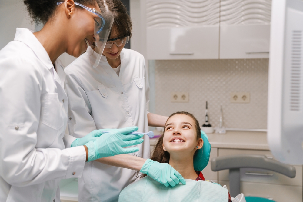  Two dentists smile at young girl seated in dental clinic chair as they prepare to examine and clean her teeth.