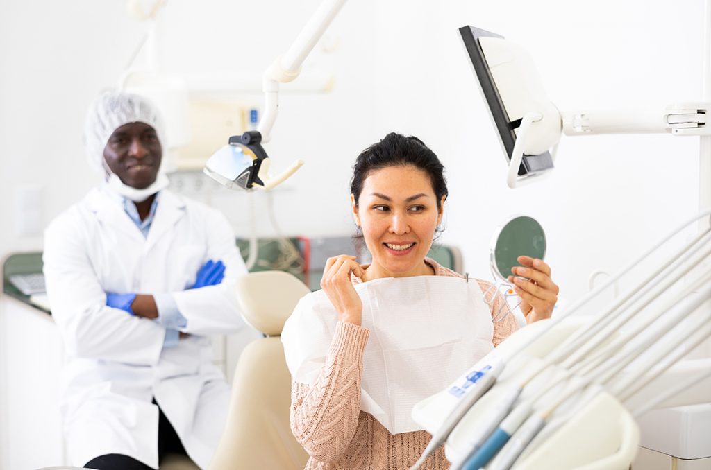  Woman in dental chair holds mirror and admires her straightened teeth as orthodontist behind watches and smiles.