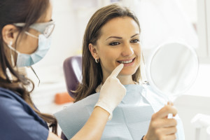  Dentist wearing mask and goggles points to dental implant in mouth of smiling woman sitting in dental chair holding a mirror. 