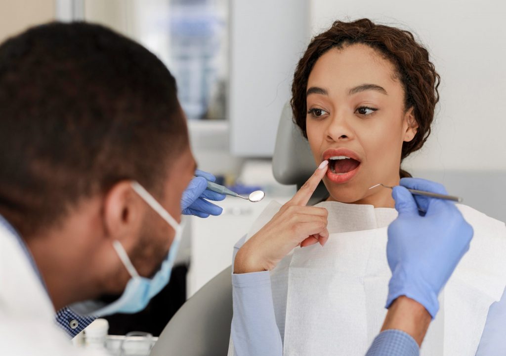 Woman in a dental chair points to her damaged tooth as if asking her dentist, “Do you know how to fix a chipped tooth?”