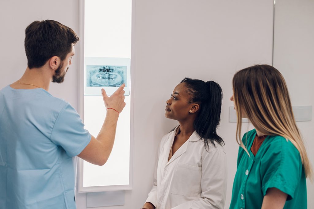 Three comprehensive dentists stand before vertical lightbox in dental clinic, examining radiograph of a patient’s teeth.