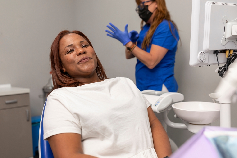  Woman reclines in a dental chair and smiles, knowing how to get over dental anxiety, as a dentist in the background puts on gloves.