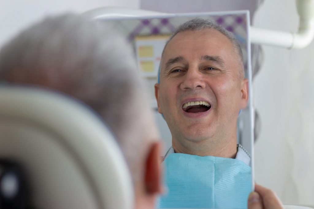 Man reclining in dental chair holds up square mirror and smiles at his reflection, showing off his affordable dental implants.