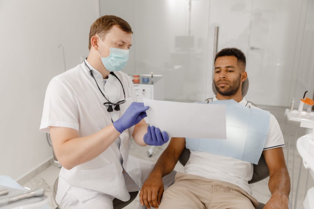 A masked dentist shows a dental X-ray to a man reclining in a dental chair, discussing a plan to repair the patient’s chipped crown.