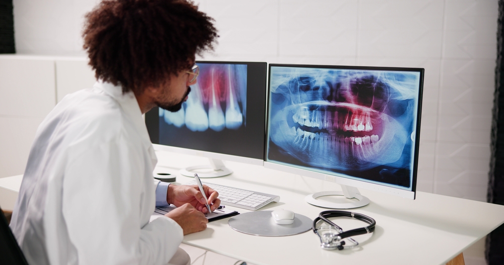  An endodontist in a medical jacket sits at a table reviewing dental X-rays that indicate a patient needs a root canal treatment. 