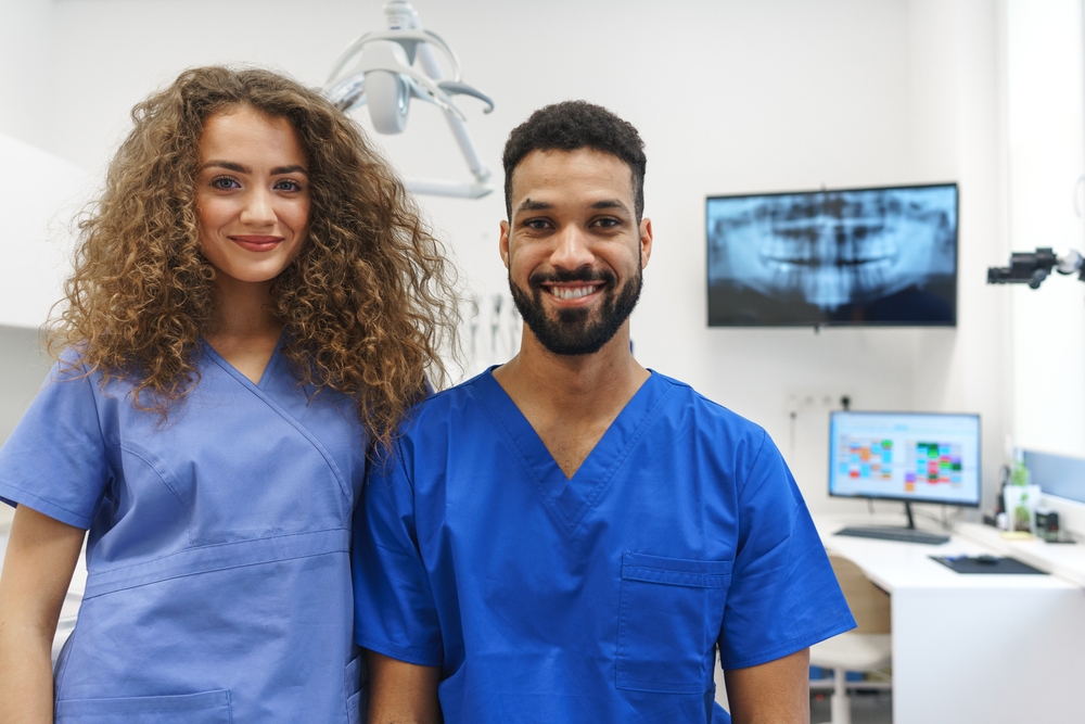  Two Philadelphia endodontists stand and smile in an office, dental X-rays visible on a wall-mounted monitor in the background. 