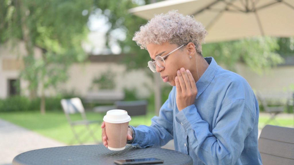 Woman sitting outside drinking coffee holds left hand to lower jaw as she feels pain from a damaged tooth.