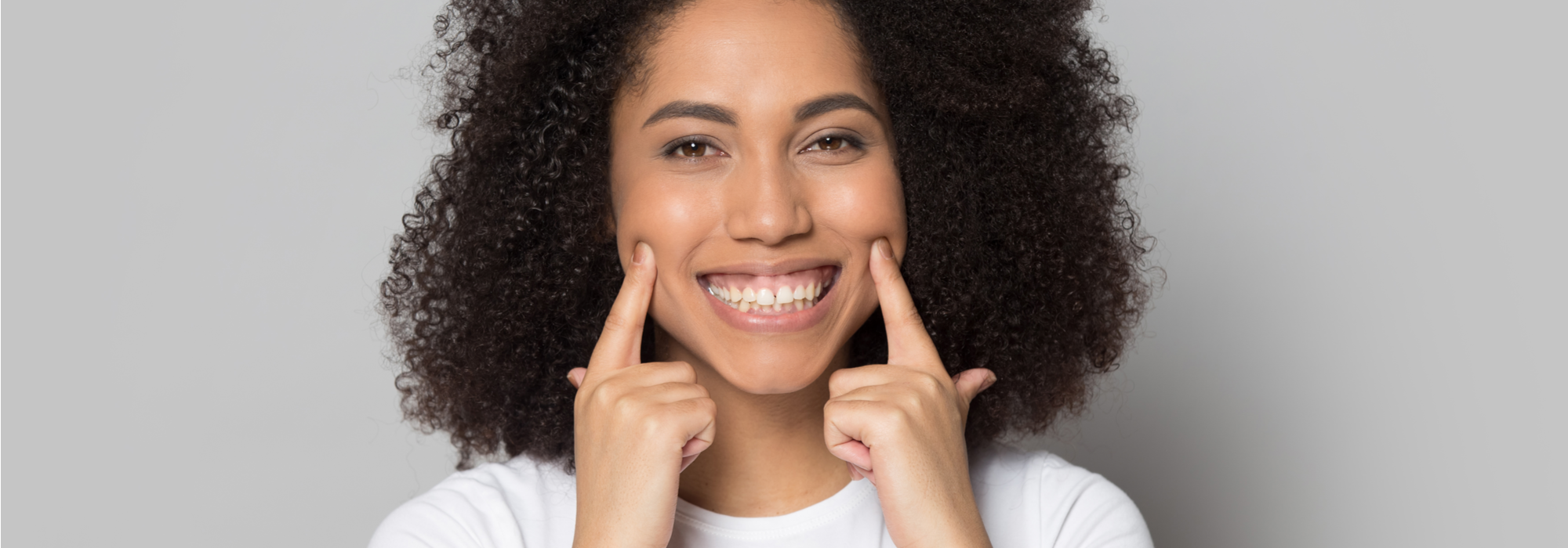 A young, lovely, African American female student with brown hair and brown eyes, wearing a white t-shirt points to her smile with both hands.