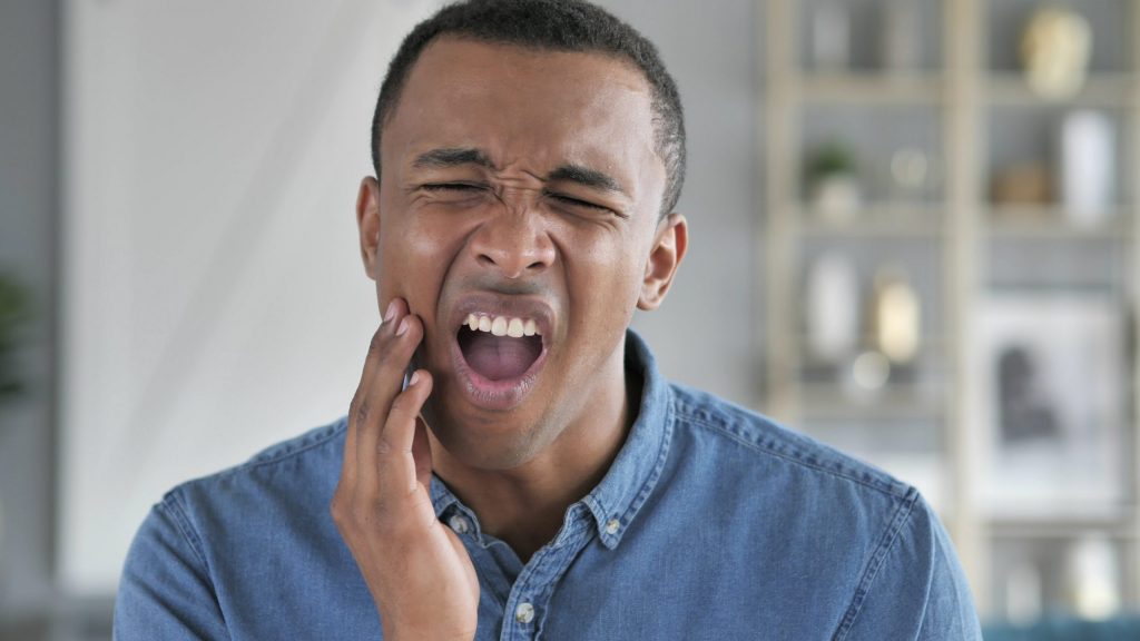 A man holds his hand to his jaw, his mouth open, suffering from a tooth infection. 