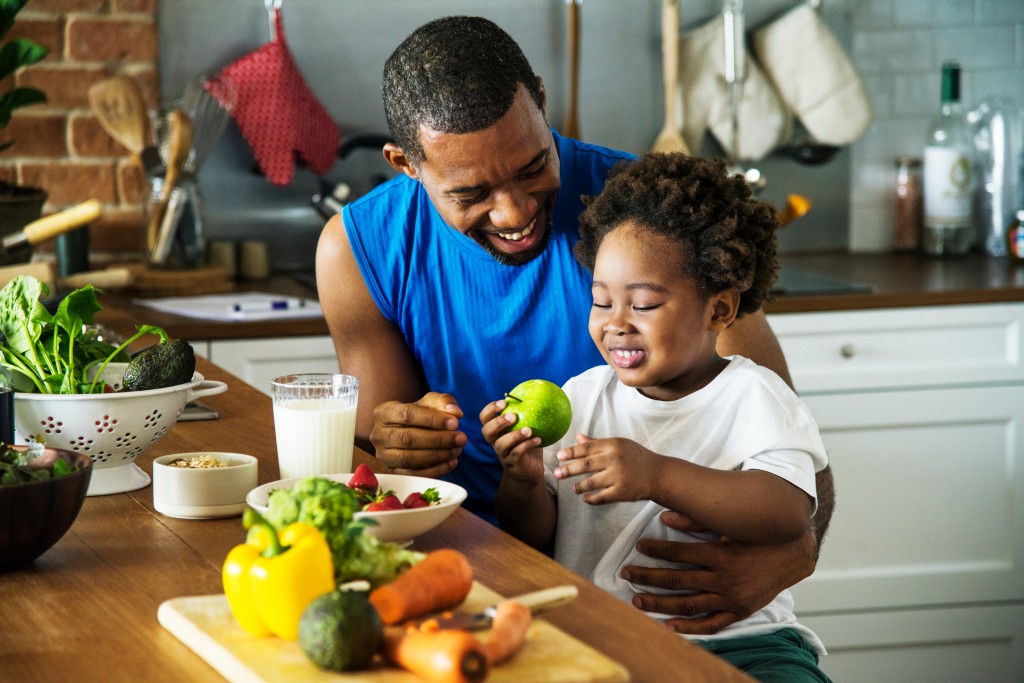 Father and toddler son cooking together in a beautiful kitchen. Son holding an apple. Fresh vegetables on a cutting board and in bowls.