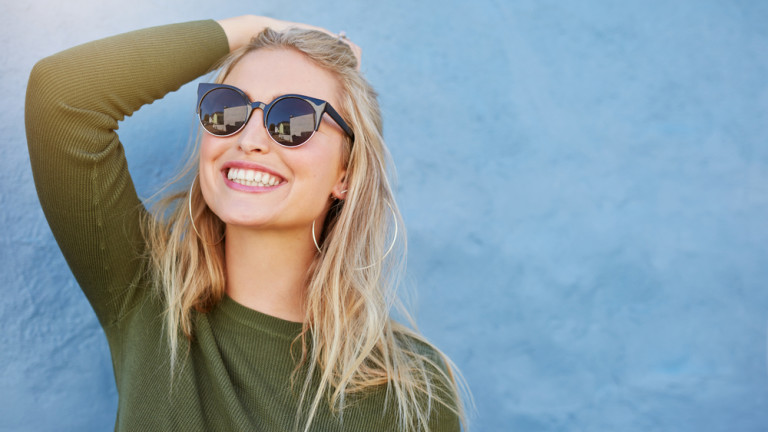 Close up shot of stylish young woman in sunglasses smiling against blue background.