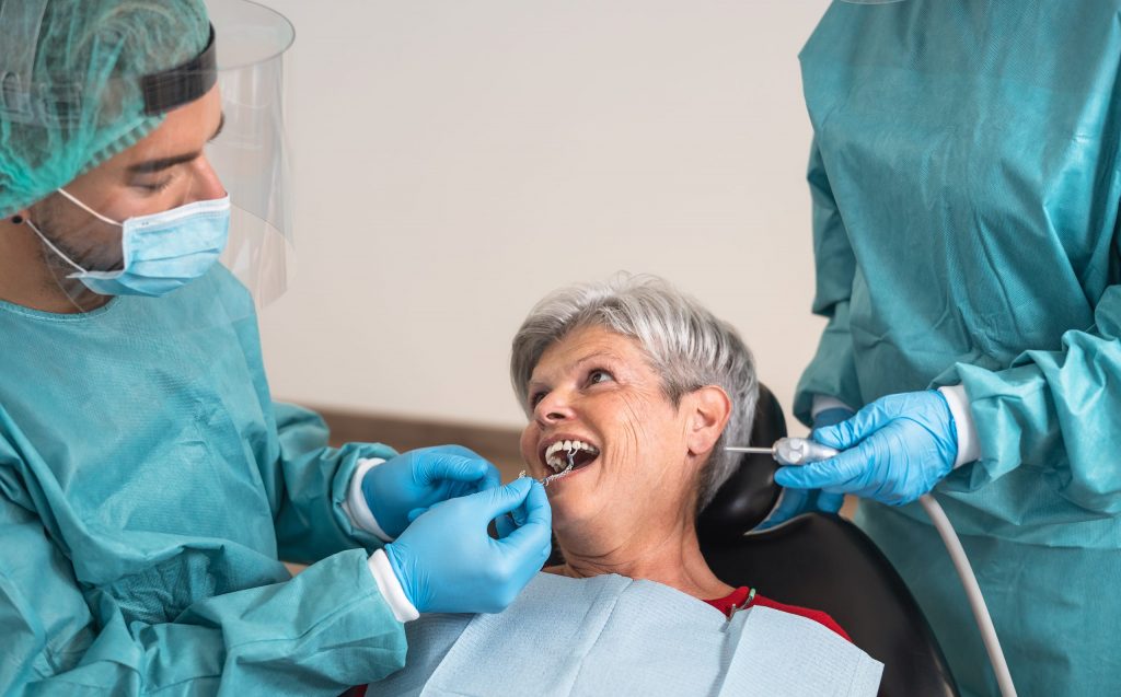 Dentists in PPE operating on an older female patient for tooth repair in a dentist’s office