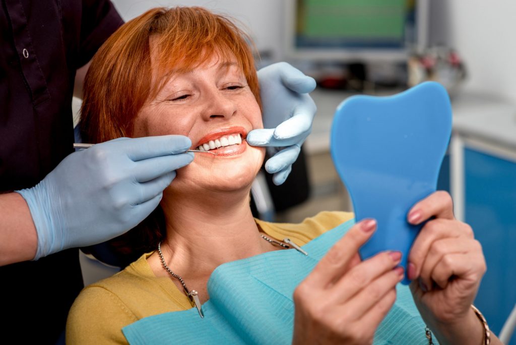 Patient sits in a dentist’s chair and smiles in mirror, displaying new dental implants, doctor in gloves assists. 