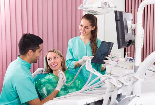A Hispanic male dental student helps treat a young woman patient, while a female dental student with long brown hair examines an X-ray.