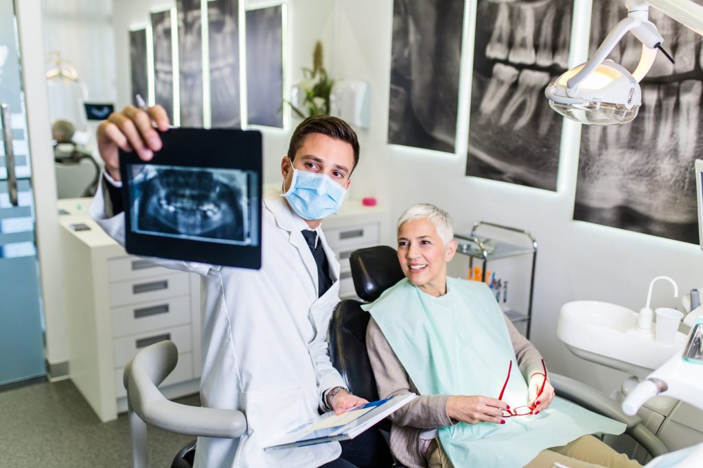  Masked Philadelphia prosthodontist reviews dental X-rays with a patient who sits in a dental examination chair. 