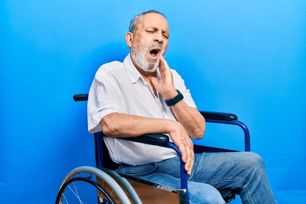 Man with disabilities in wheelchair shuts his eyes and holds left hand to jaw, indicating he feels oral health care.