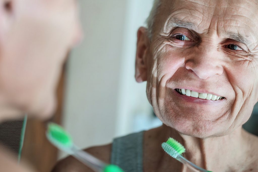 Elderly man smiles in the mirror while brushing his teeth