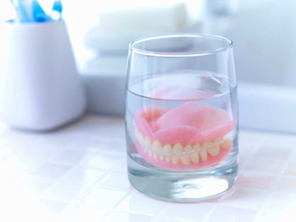  Dentures soaking in a glass on the bathroom counter with toothbrushes in the background.