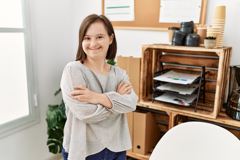 Woman with Down syndrome stands in her office with arms crossed, smiling after having had a successful dental treatment.
