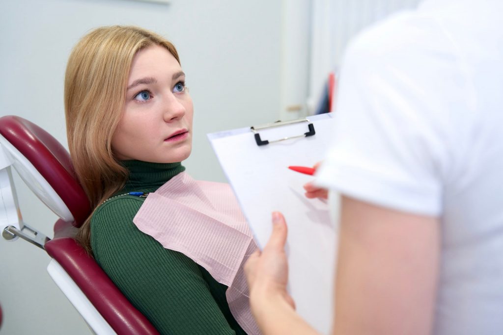  Teenage girl reclines in dental chair, looking at her dentist with wide eyes, illustrating need for dental anxiety management. 