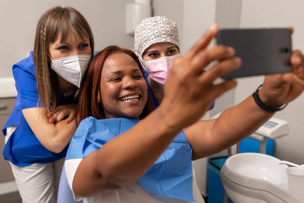 Smiling woman in dental chair takes selfie with her two masked dentists, no longer feeling dental phobia and anxiety. 