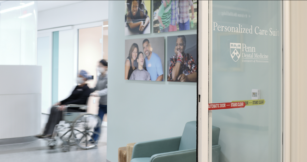  Dentist at Penn Dental Medicine pushes patient with MS symptoms in wheelchair down a hall in the Personalized Care Suite.