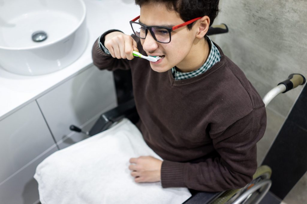  Teenager with MS in wheelchair sits next to a sink, brushing his teeth, important for good oral hygiene. 