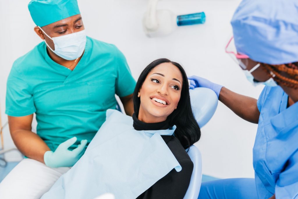 A woman reclines in a dental chair, talking with a dentist and student dentist at the clinic of a dental school that takes patients.