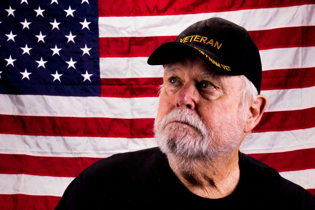 An older vet stands in front of a U.S. flag in a black T-shirt and Vietnam Veterans hat, and wonders where to find a dentist for veterans. 