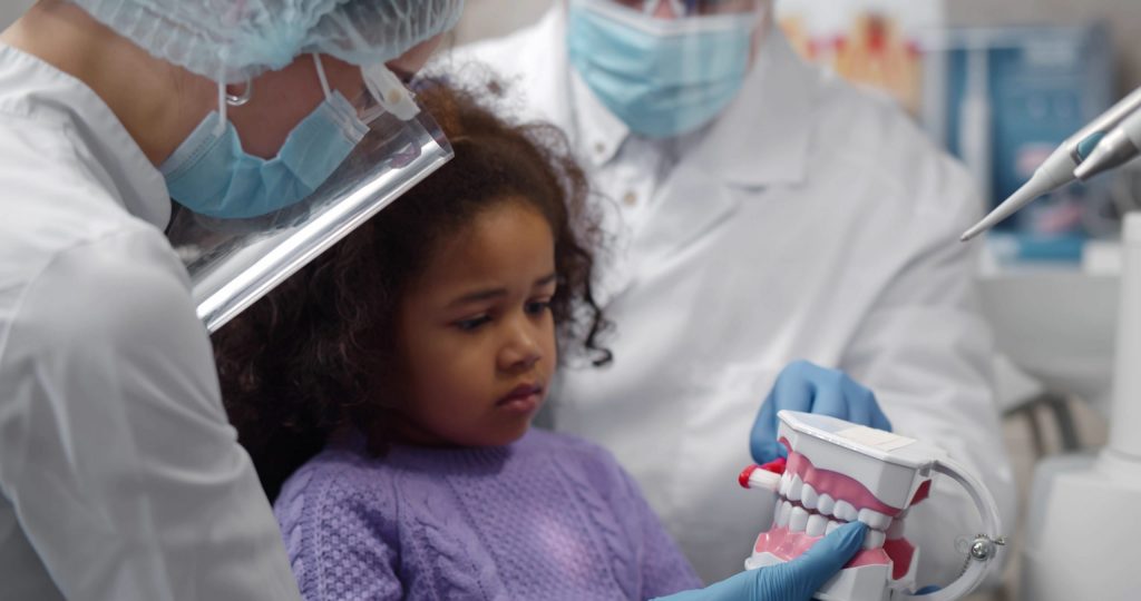  Two dentists wearing PPE show young girl in dental chair model of teeth, trying to help allay her pediatric dentist anxiety.