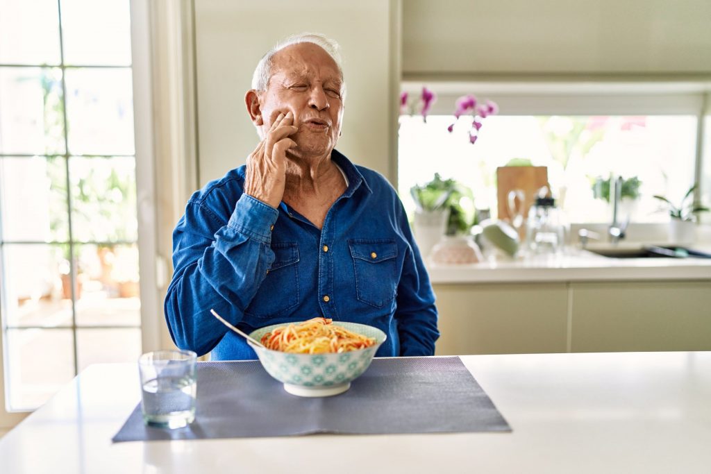 Man sits at his kitchen counter with a bowl of pasta, pressing his right hand to his cheek due to pain from mouth sores.
