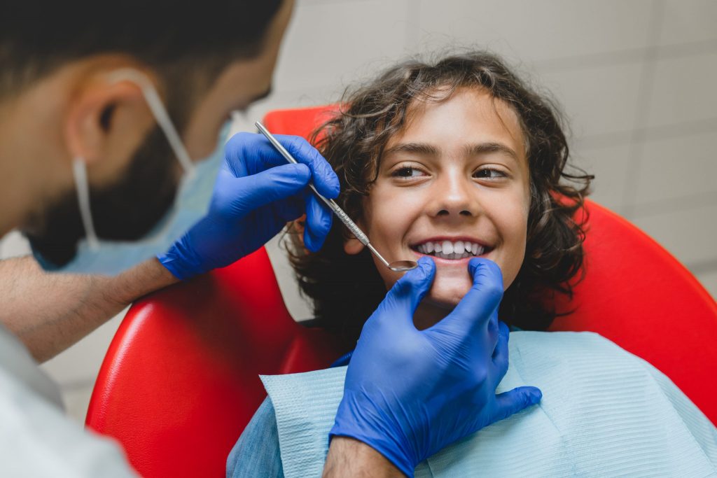 Teenage boy sits in dental chair, feeling no dental anxiety and smiling at dentist as dentist holds dental tool to his mouth.