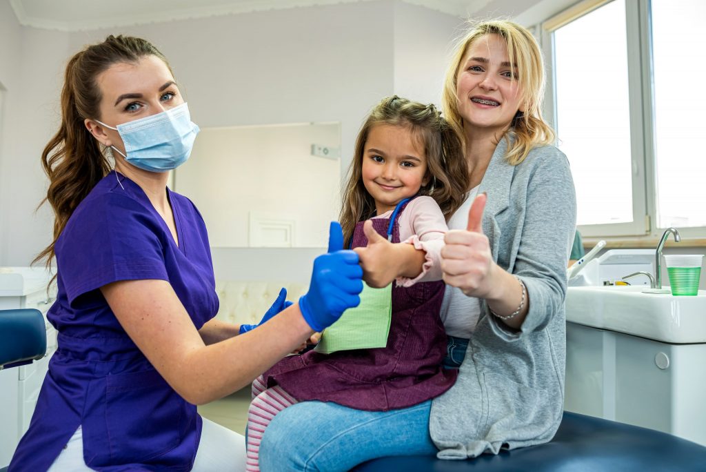  Young girl sits in mother’s lap at dentist’s office, both of them and dentist making “thumbs-up” sign to show no dental anxiety.