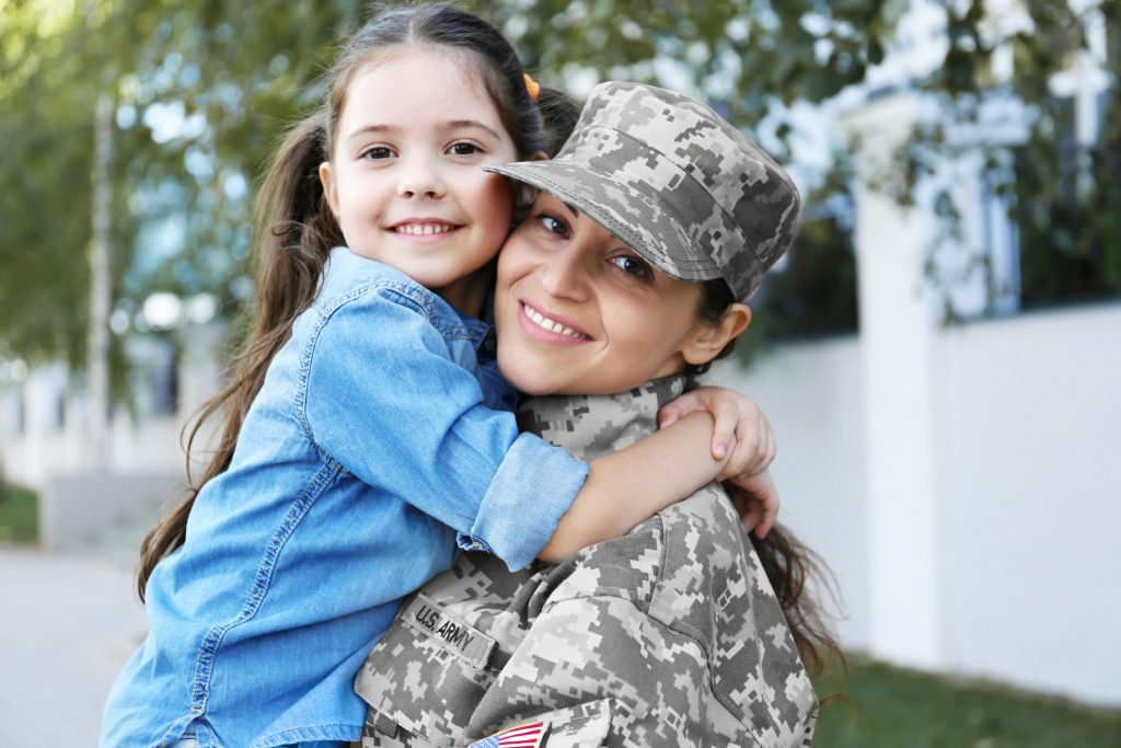  Female vet in fatigues holds her daughter outside and smiles because she has dental care for veterans without insurance at PDM.