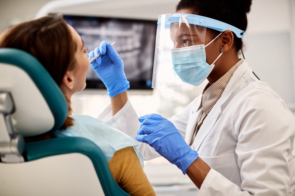 An endodontist wearing PPE examines the teeth of a woman, reclining in a dental chair, who complains of signs of tooth infection.