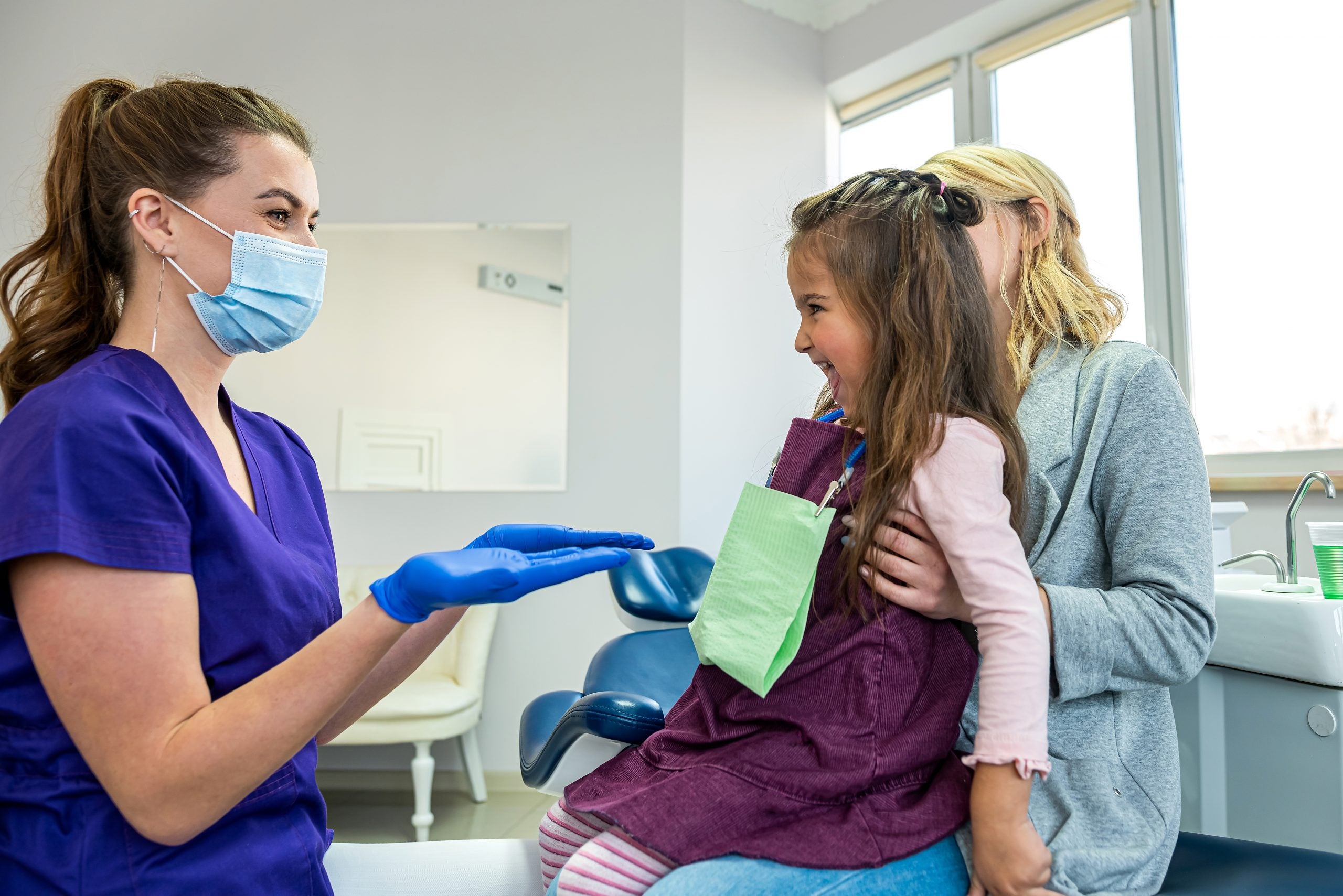 Pediatric dentist wearing face mask and gloves holds hands out to smiling young girl wearing dental bib sitting on mother’s lap.