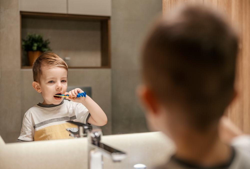 Toddler stands at bathroom sink in front of mirror, practicing brushing his teeth.