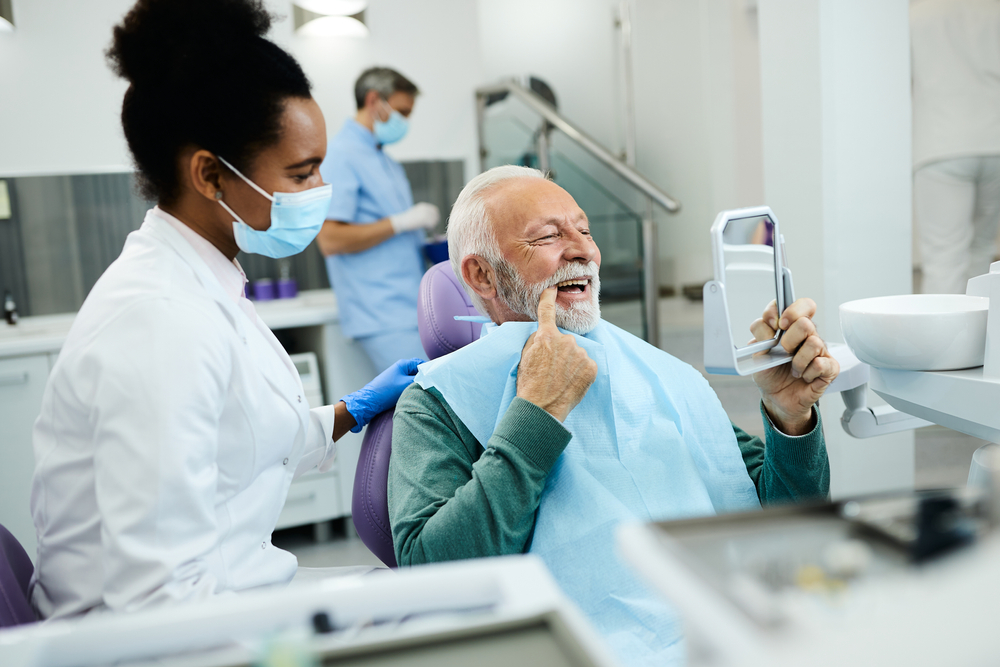 A dentist, wearing a mask, watches her patient in a chair look at his teeth in a mirror after dental treatment.