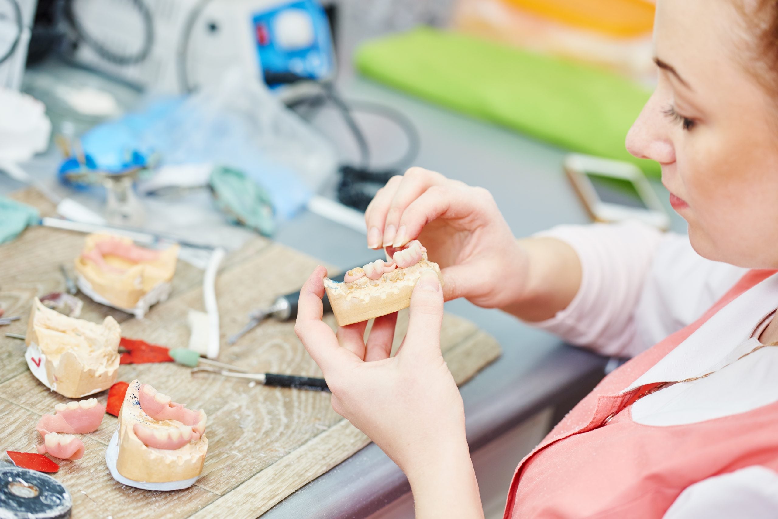 A dental lab technician sits at a workstation, forming dentures to fit a mold of teeth, with finished sets of dentures in front of her.
