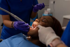 A Black woman reclines in a dental chair while gloved dentists provide low-income dental care.