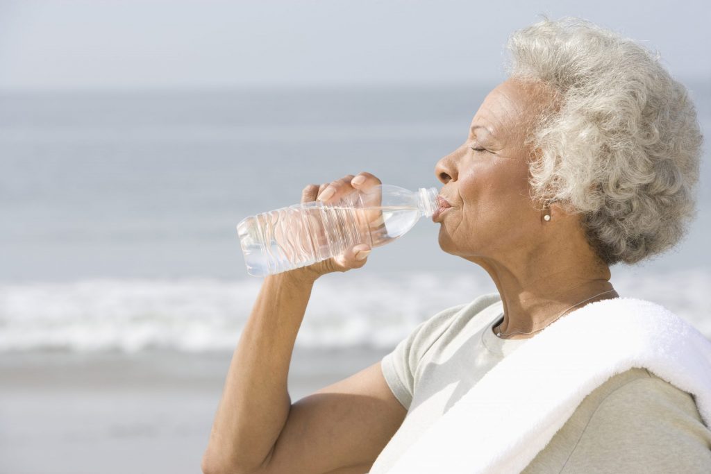 A woman stands on a beach, ocean in the background, sipping water from a plastic bottle to help alleviate her dry mouth symptoms.