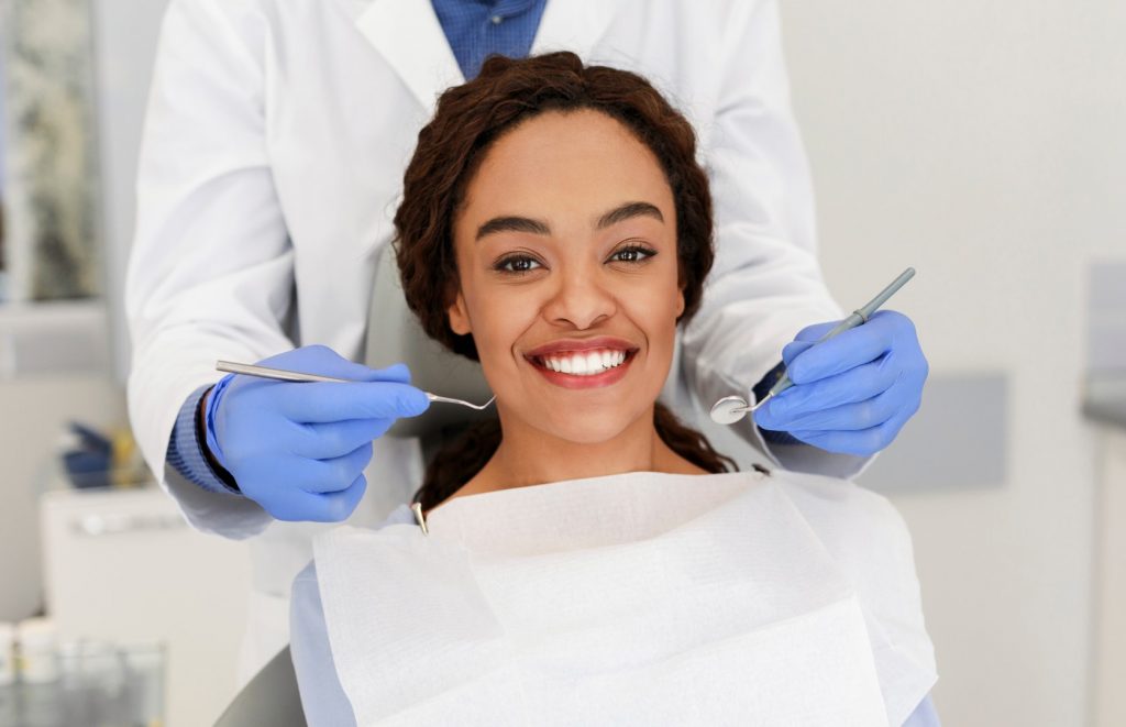 A young woman smiles while a dentist wearing gloves stands behind her holding tools for an exam.