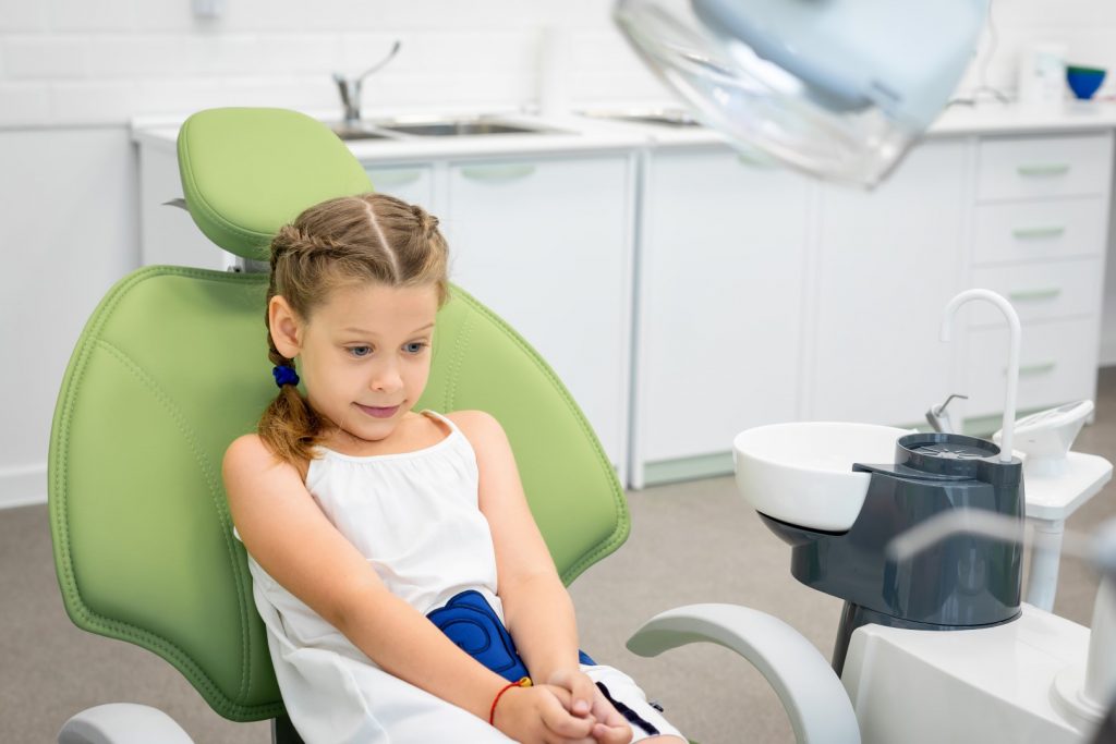 A young elementary school-age girl sits in a dental chair looking worried, illustrating dental anxiety in children.